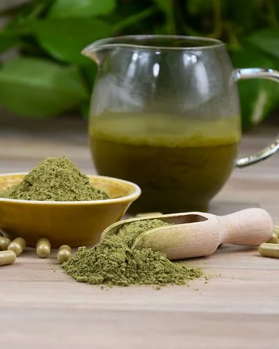 Auf einer Holzplatte befinden sich eine Glaskanne mit grünem Aufguss sowie eine Keramikschale, eine hölzerne Apothekerschaufel und zahlreiche transparente Kapseln, die jeweils mit grünem Pulver gefüllt sind.