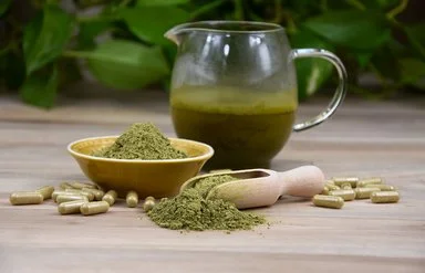 Auf einer Holzplatte befinden sich eine Glaskanne mit grünem Aufguss sowie eine Keramikschale, eine hölzerne Apothekerschaufel und zahlreiche transparente Kapseln, die jeweils mit grünem Pulver gefüllt sind.