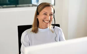 Frau sitzt mit Headset vor einem Computer und lächelt. 