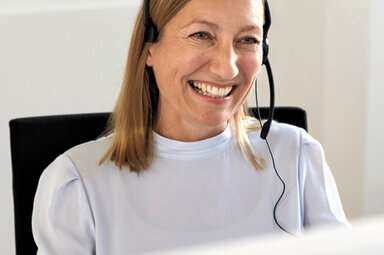 Frau sitzt mit Headset vor einem Computer und lächelt. 
