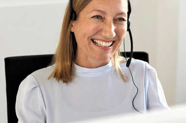 Frau sitzt mit Headset vor einem Computer und lächelt.