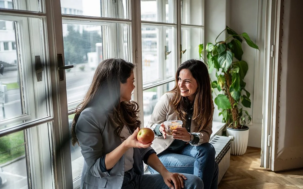 Zwei Frauen sitzen am Fenster und machen Pause. Eine Frau hält einen Apfel in der Hand, die andere eine Schale mit Gemüsesalat.