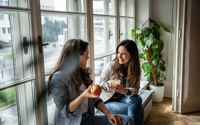Zwei Frauen sitzen am Fenster und machen Pause. Eine Frau hält einen Apfel in der Hand, die andere eine Schale mit Gemüsesalat.