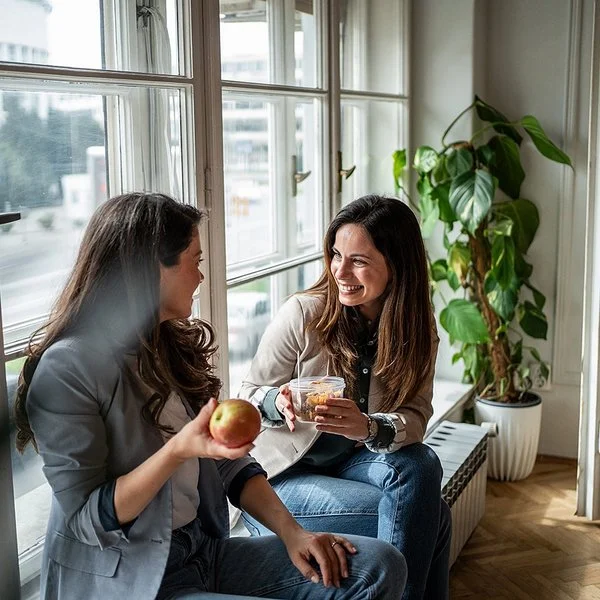 Zwei Frauen sitzen am Fenster und machen Pause. Eine Frau hält einen Apfel in der Hand, die andere eine Schale mit Gemüsesalat.