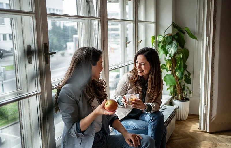 Zwei Frauen sitzen am Fenster und machen Pause. Eine Frau hält einen Apfel in der Hand, die andere eine Schale mit Gemüsesalat.