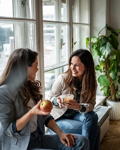 Zwei Frauen sitzen am Fenster und machen Pause. Eine Frau hält einen Apfel in der Hand, die andere eine Schale mit Gemüsesalat.