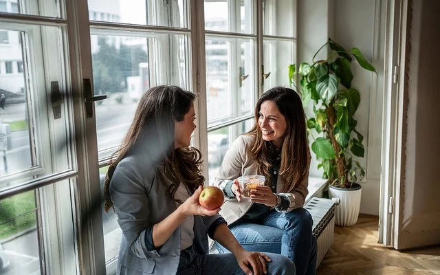 Zwei Frauen sitzen am Fenster und machen Pause. Eine Frau hält einen Apfel in der Hand, die andere eine Schale mit Gemüsesalat.