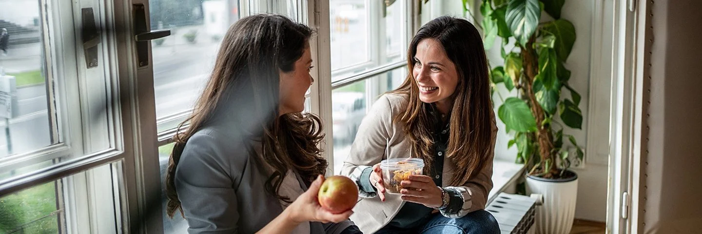 Zwei Frauen sitzen am Fenster und machen Pause. Eine Frau hält einen Apfel in der Hand, die andere eine Schale mit Gemüsesalat.