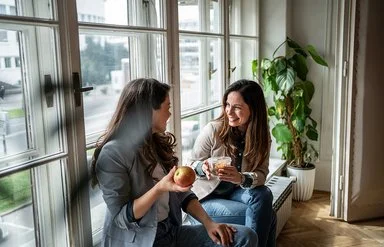 Zwei Frauen sitzen am Fenster und machen Pause. Eine Frau hält einen Apfel in der Hand, die andere eine Schale mit Gemüsesalat.