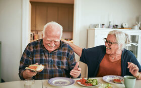 Zwei Senioren sitzen nebeneinander am Küchentisch und genießen lachend ihre gesunde Ernährung.
