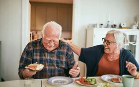 Zwei Senioren sitzen nebeneinander am Küchentisch und genießen lachend ihre gesunde Ernährung.
