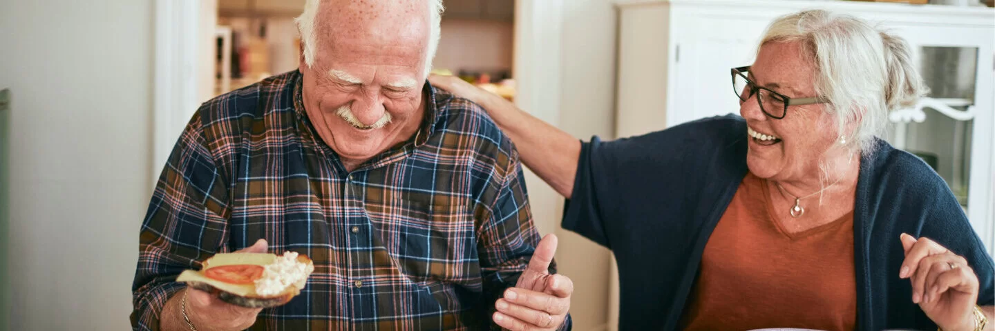 Zwei Senioren sitzen nebeneinander am Küchentisch und genießen lachend ihre gesunde Ernährung.