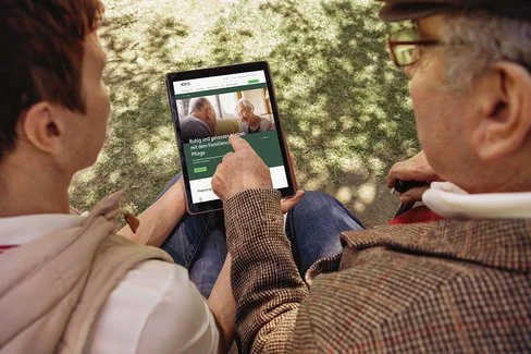 Ein älterer und ein jüngerer Mann lesen Texte auf einem Tablet-Computer. Die AOK informiert mit gedruckten Magazinen und auch digital.