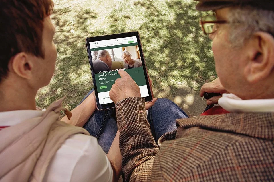 Ein älterer und ein jüngerer Mann lesen Texte auf einem Tablet-Computer. Die AOK informiert mit gedruckten Magazinen und auch digital.