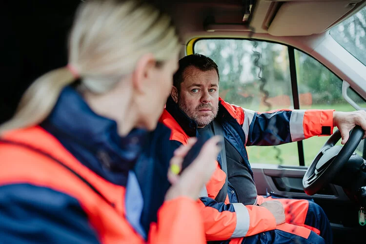 Ein Mann sitzt am Steuer eines Rettungswagens und schaut zur Beifahrerin herüber, die ein Funkgerät in der Hand hält.