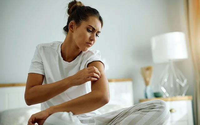 Eine junge Frau im Schlafanzug sitzt auf Ihrem Bett und begutachtet mit kritischem Blick die Haut an ihrem Oberarm.