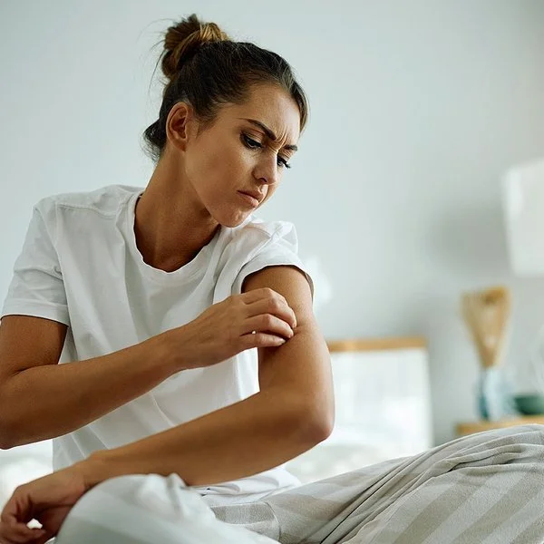 Eine junge Frau im Schlafanzug sitzt auf Ihrem Bett und begutachtet mit kritischem Blick die Haut an ihrem Oberarm.