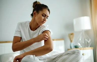 Eine junge Frau im Schlafanzug sitzt auf Ihrem Bett und begutachtet mit kritischem Blick die Haut an ihrem Oberarm.