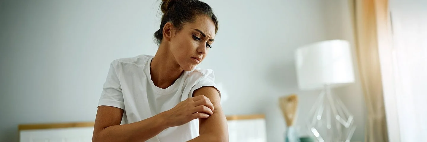 Eine junge Frau im Schlafanzug sitzt auf Ihrem Bett und begutachtet mit kritischem Blick die Haut an ihrem Oberarm.