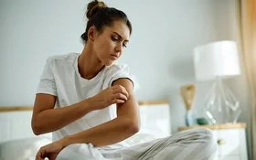 Eine junge Frau im Schlafanzug sitzt auf Ihrem Bett und begutachtet mit kritischem Blick die Haut an ihrem Oberarm.