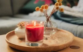 Eine rote Duftkerze im Glas, neben einer Porzellandose und einem kleinen Strauß Trockenblumen auf einem hölzernen Tablett.