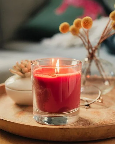 Eine rote Duftkerze im Glas, neben einer Porzellandose und einem kleinen Strauß Trockenblumen auf einem hölzernen Tablett.