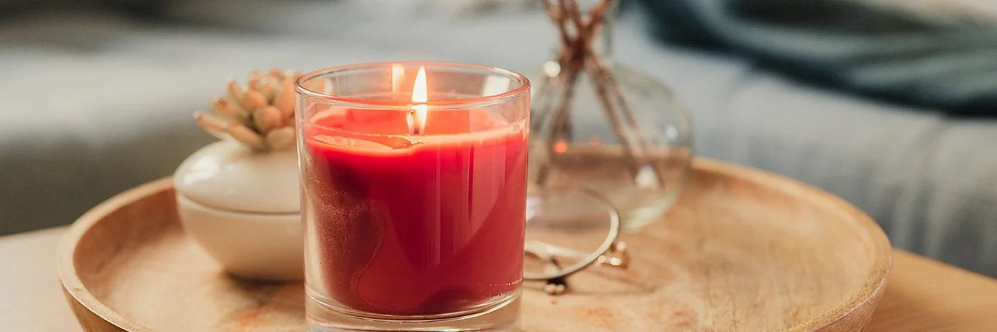 Eine rote Duftkerze im Glas, neben einer Porzellandose und einem kleinen Strauß Trockenblumen auf einem hölzernen Tablett.