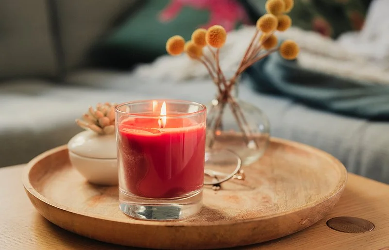 Eine rote Duftkerze im Glas, neben einer Porzellandose und einem kleinen Strauß Trockenblumen auf einem hölzernen Tablett.