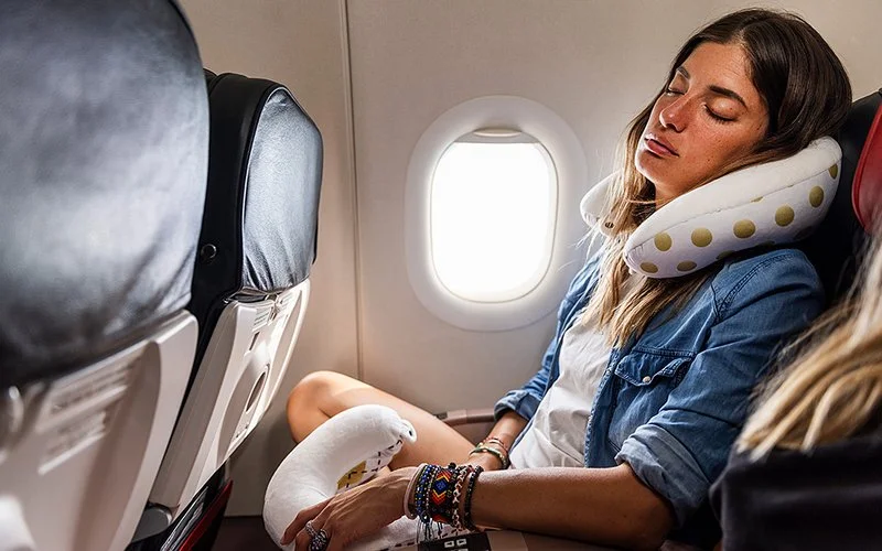 Eine junge Frau schläft in ihrem Flugzeugsitz am Fenster, mit einem Nackenkissen um den Hals. Sie ist sommerlich gekleidet.