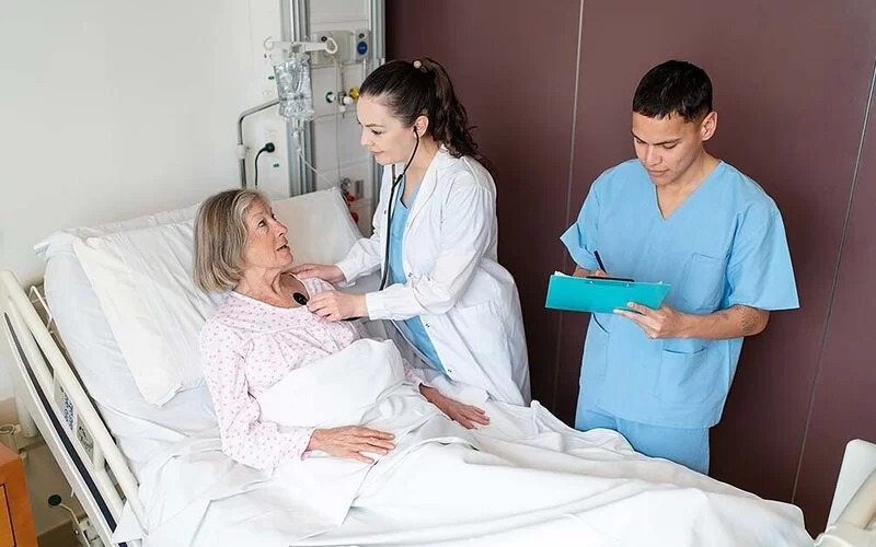 Ein Zimmer in einem Krankenhaus. Eine Ärztin horcht eine im Bett liegende Patientin mit einem Stethoskop ab. Ein Pfleger steht neben der Ärztin und macht Notizen.