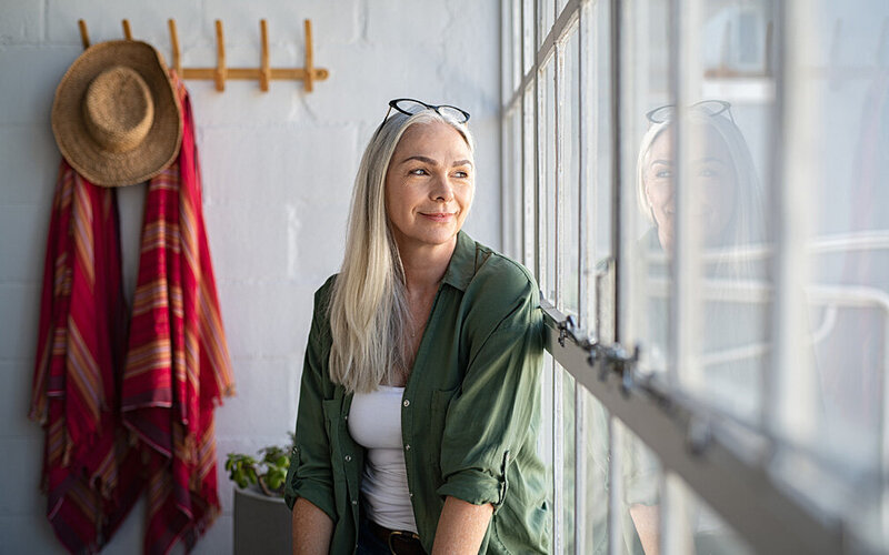 Frau mit langen grauen Haaren, vermutlich in den Wechseljahren, sitzt versonnen am Fenster und guckt raus.