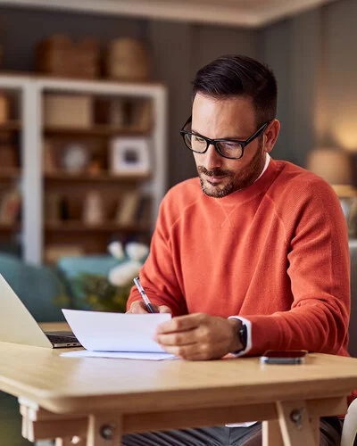 Ein Mann in einem orangefarbenen Pullover sitzt an einem Schreibtisch in häuslicher Umgebung und macht Notizen.