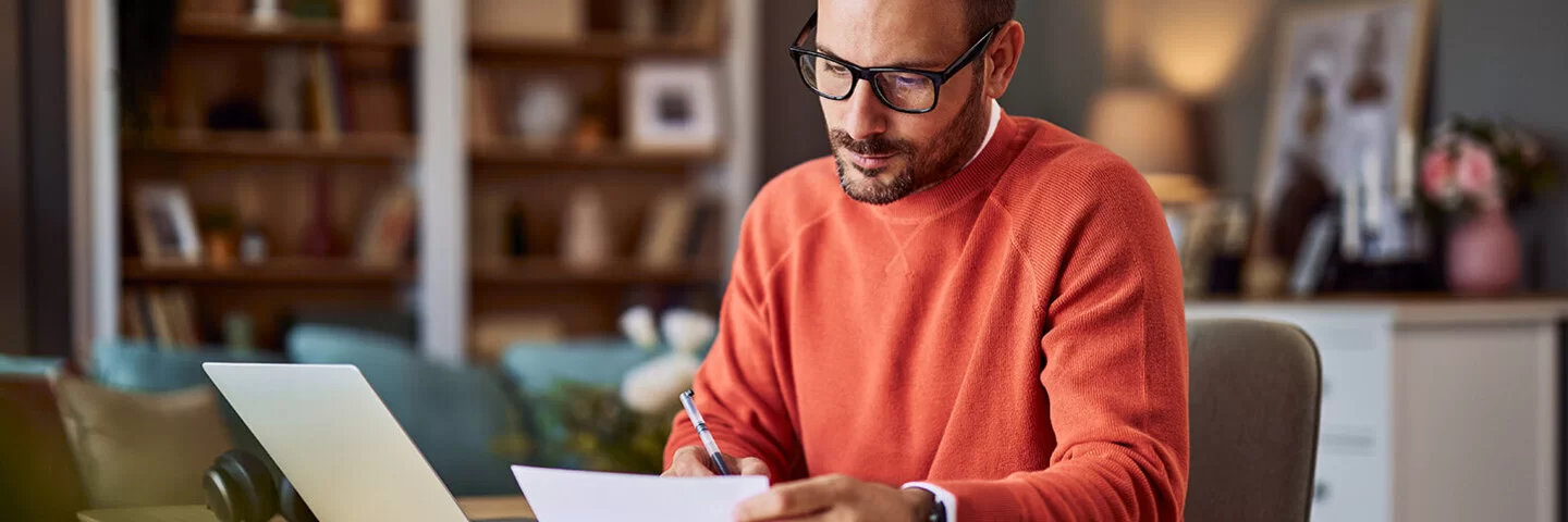 Ein Mann in einem orangefarbenen Pullover sitzt an einem Schreibtisch in häuslicher Umgebung und macht Notizen.