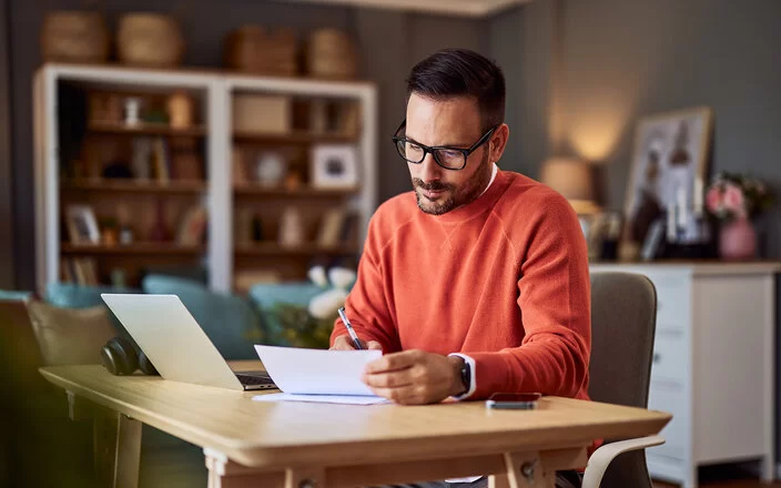 Ein Mann in einem orangefarbenen Pullover sitzt an einem Schreibtisch in häuslicher Umgebung und macht Notizen.