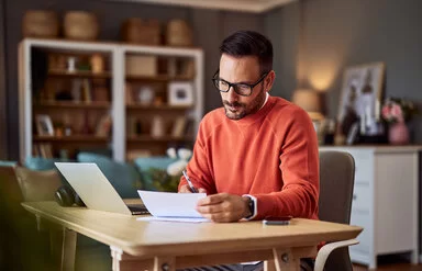 Ein Mann in einem orangefarbenen Pullover sitzt an einem Schreibtisch in häuslicher Umgebung und macht Notizen.