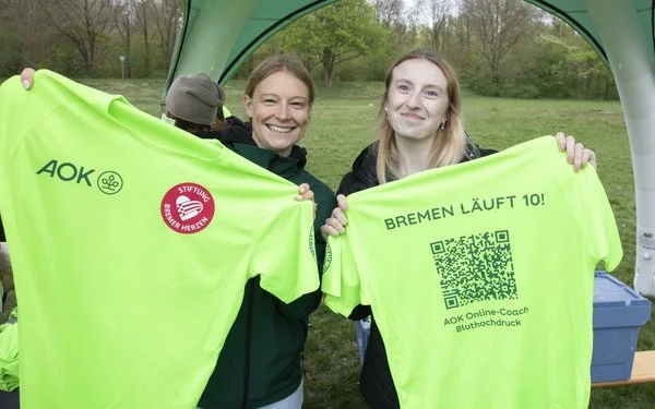 Zwei Sportlerinnen halten ein Trikot in leuchtendem Grün mit AOK-Logo und Stiftung Bremer Herzen hoch.