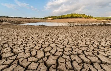 Eine Landschaftsaufnahme zeigt einen ausgetrockneten, rissigen Boden. Im Hintergrund befinden sich ein Teich mit niedrigem Wasserstand und eine kleine bewaldete Anhöhe.