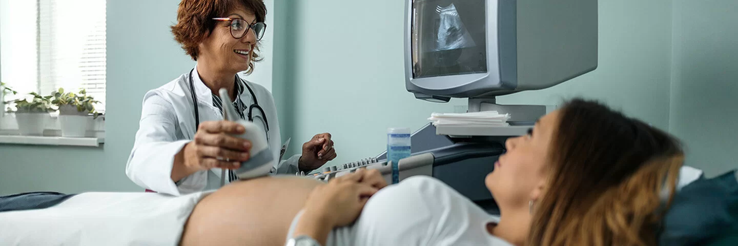 Eine lächelnde Ärztin führt bei einer schwangeren Frau eine Ultraschalluntersuchung des Bauches durch. Das Ultraschallbild ist auf einem Bildschirm über dem Bauch der Frau zu sehen.