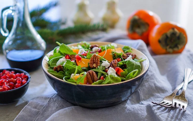 Eine Schüssel mit grünem Salat steht auf einem Tisch, darin auch Kakistücke und Walnüsse. Daneben liegen ganze Kakis.