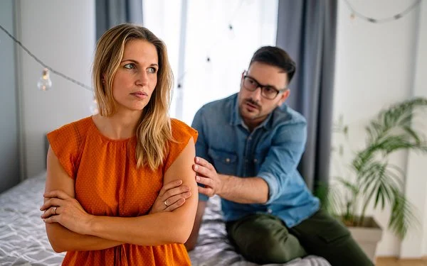 Eine junge Frau sitzt mit verschränkten Armen auf einem Bett. Sie wirkt uneinsichtig. Ein Mann redet im Hintergrund auf sie ein.