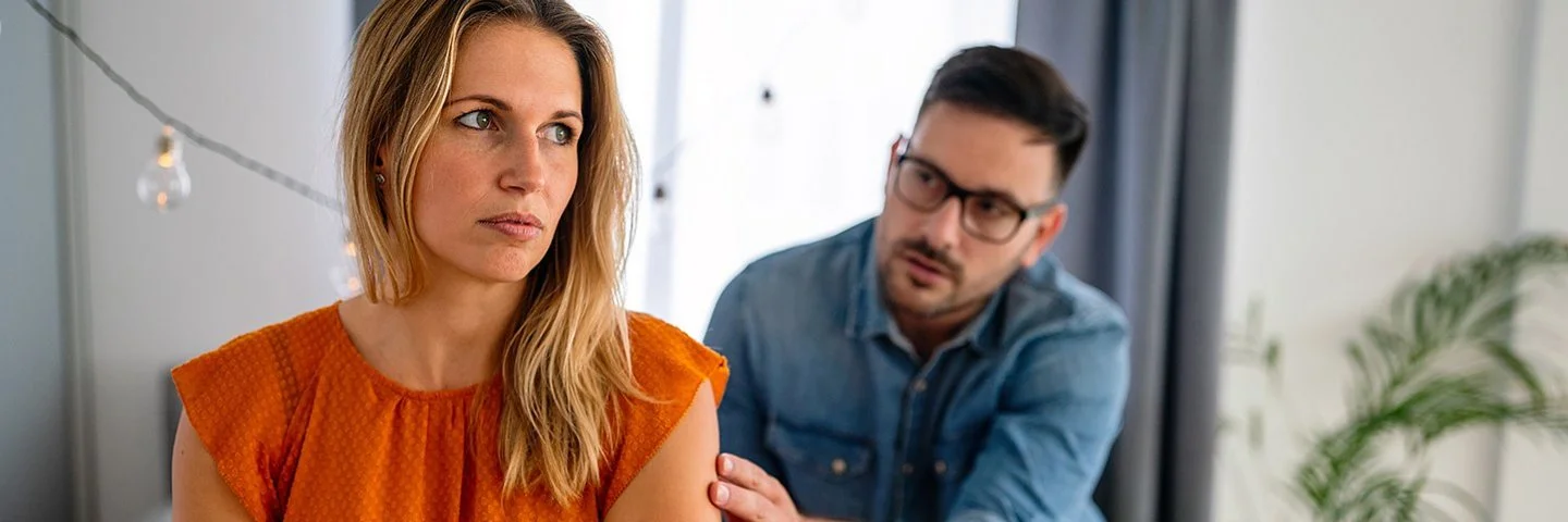 Eine junge Frau sitzt mit verschränkten Armen auf einem Bett. Sie wirkt uneinsichtig. Ein Mann redet im Hintergrund auf sie ein.