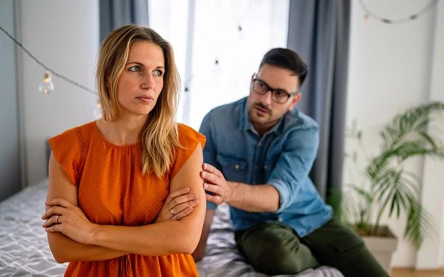 Eine junge Frau sitzt mit verschränkten Armen auf einem Bett. Sie wirkt uneinsichtig. Ein Mann redet im Hintergrund auf sie ein.