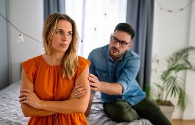 Eine junge Frau sitzt mit verschränkten Armen auf einem Bett. Sie wirkt uneinsichtig. Ein Mann redet im Hintergrund auf sie ein.