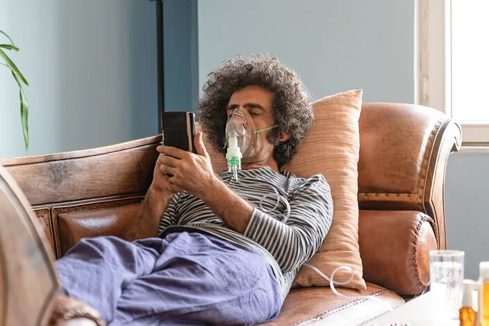Ein Mann liegt auf einem Sofa und trägt ein Beatmungsgerät, während er auf ein Handy in seiner Hand schaut.