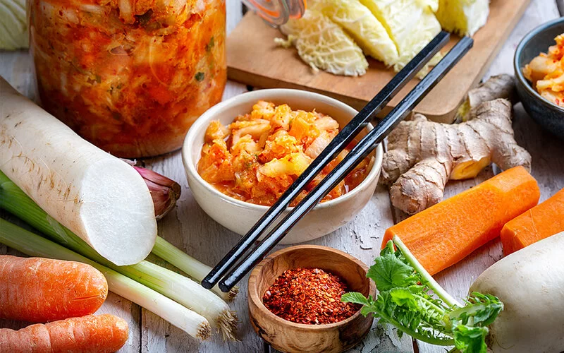Ein Glas mit Kimchi steht auf einem hölzernen Untergrund, daneben liegen verschiedene Gemüsesorten.