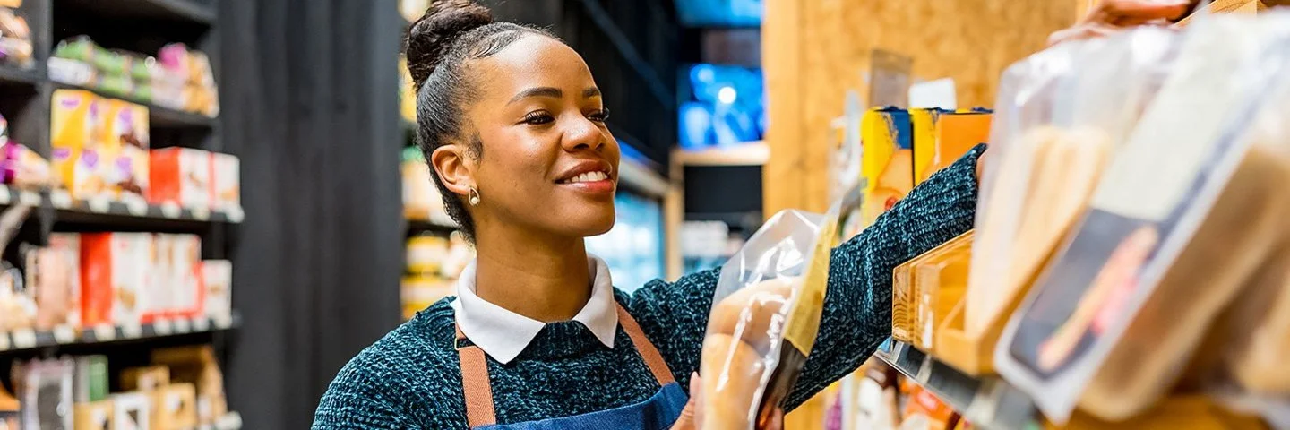 Eine Angestellte in einem Supermarkt sortiert in Folie verpackte Brotwaren in ein Regal.