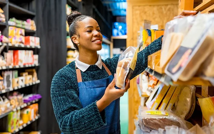 Eine Angestellte in einem Supermarkt sortiert in Folie verpackte Brotwaren in ein Regal.
