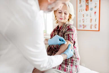  Eine Frau sitzt in einem Behandlungszimmer auf einer Behandlungsliege. Ein Arzt desinfiziert ihren Oberarm, um eine Impfung vorzubereiten.