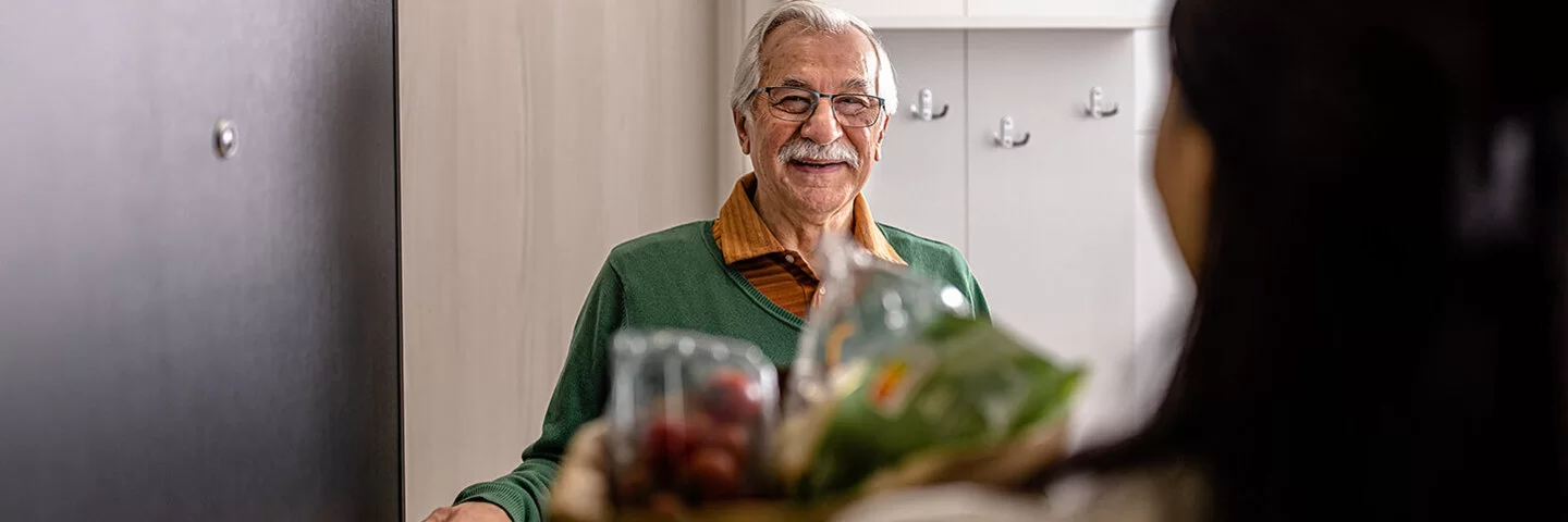 Ein älterer Herr öffnet einer Frau, die ihn besucht die Türe und lacht.