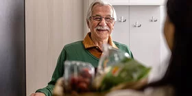 Ein älterer Herr öffnet einer Frau, die ihn besucht die Türe und lacht.